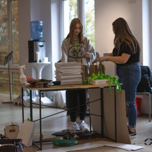 Dwie kobiety (wolontariuszki) układające na stole identyfikatory i katalogi dotyczące Festiwalu.