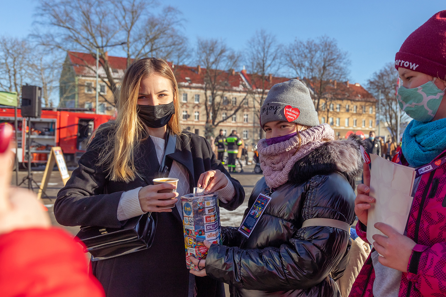 Dwie dziewczyny w kurtkach zimowych, dziewczyna po prawej stronie ma czarną maseczkę na twarzy, w lewej ręce trzyma kubek papierowy, a prawą ręką wrzuca pieniądze do skarbonki WOŚP którą trzyma dziewczynka po lewej stronie.
